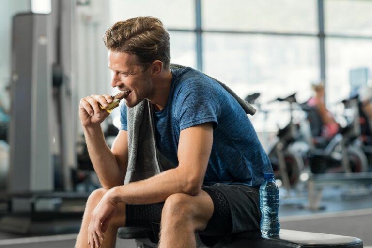 An athletic man eating a No Cow protein bar while sitting on a workout bench at the gym